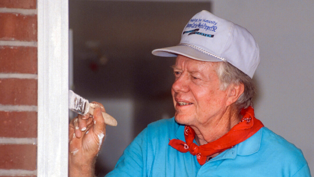 Former President Jimmy Carter, wearing a white Habitat for Humanity baseball cap, turquoise shirt, and red bandana, smiles while painting a door frame during a 1992 home-building project. His paint-covered hands hold a brush.