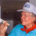 Former President Jimmy Carter, wearing a white Habitat for Humanity baseball cap, turquoise shirt, and red bandana, smiles while painting a door frame during a 1992 home-building project. His paint-covered hands hold a brush.