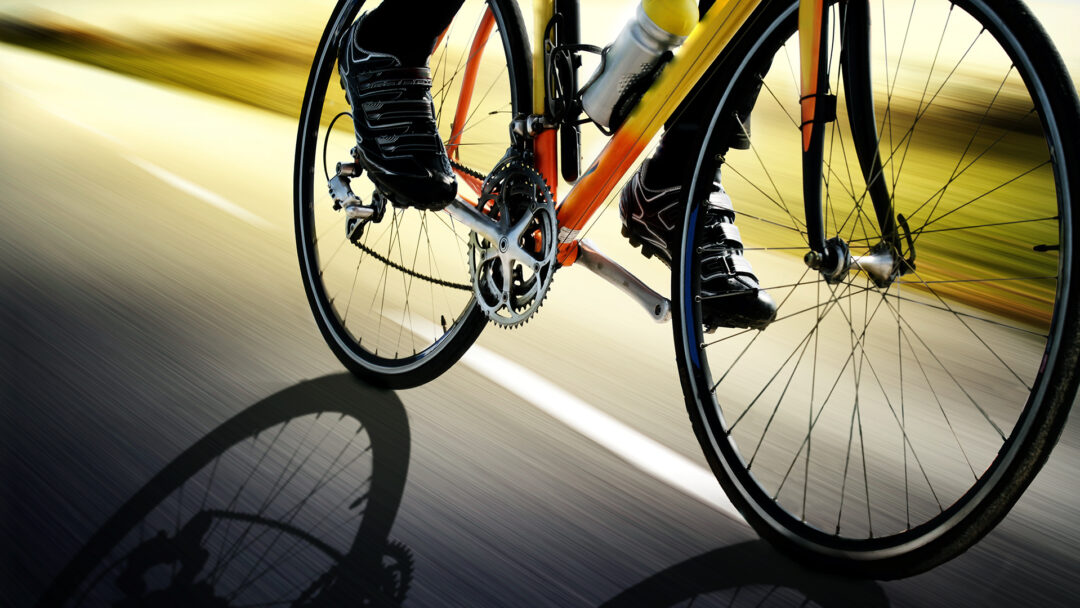 Close-up shot of a road bike in motion, showing the wheels, chainring, and pedals with black cycling shoes. The image has a dynamic motion blur effect with a yellow-tinted background. The bike appears to be orange or red in color, with black tires and a water bottle mounted on the frame. A dramatic shadow of the bicycle wheel is cast on the ground beneath.