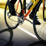 Close-up shot of a road bike in motion, showing the wheels, chainring, and pedals with black cycling shoes. The image has a dynamic motion blur effect with a yellow-tinted background. The bike appears to be orange or red in color, with black tires and a water bottle mounted on the frame. A dramatic shadow of the bicycle wheel is cast on the ground beneath.