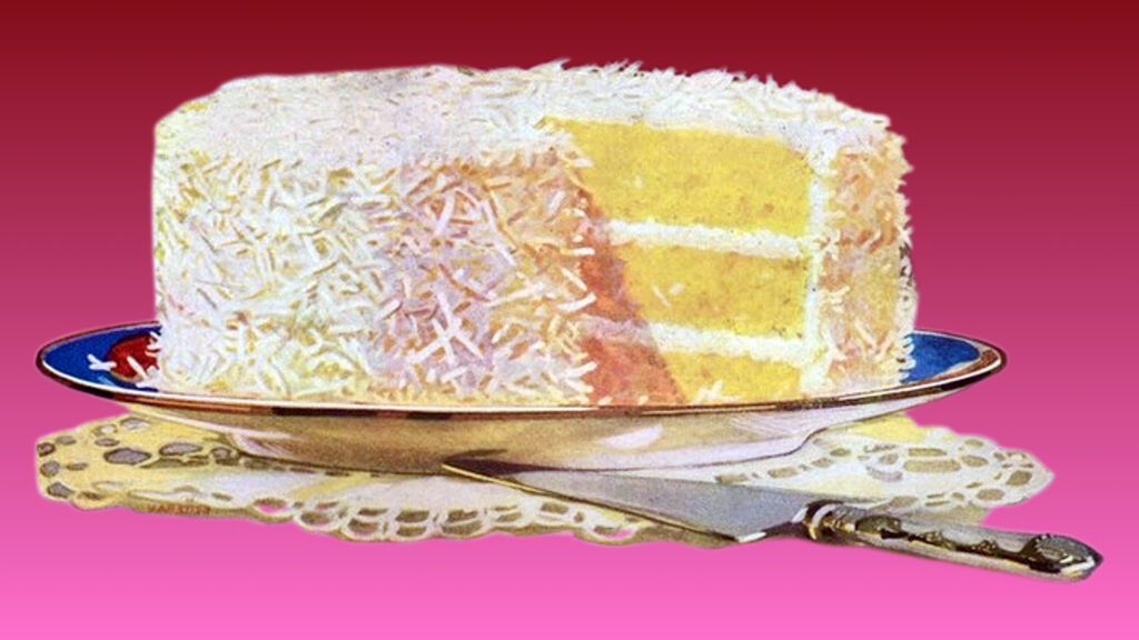 A layered coconut cake with white frosting on a blue-rimmed plate, served on a decorative doily with a silver cake server. The cake is completely covered in shredded coconut and is three layers tall.