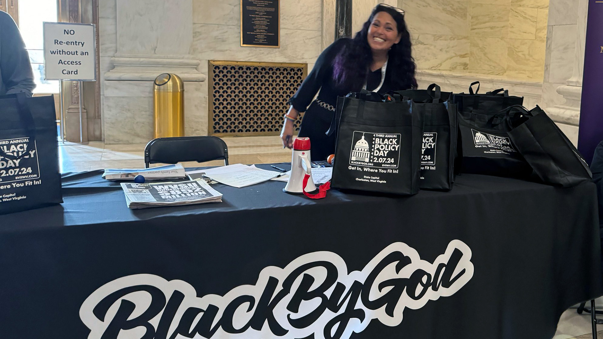 A smiling woman staffs the Black By God information table at Black Policy Day 2024, with promotional tote bags and materials displayed on a black tablecloth featuring the organization's bold white logo.