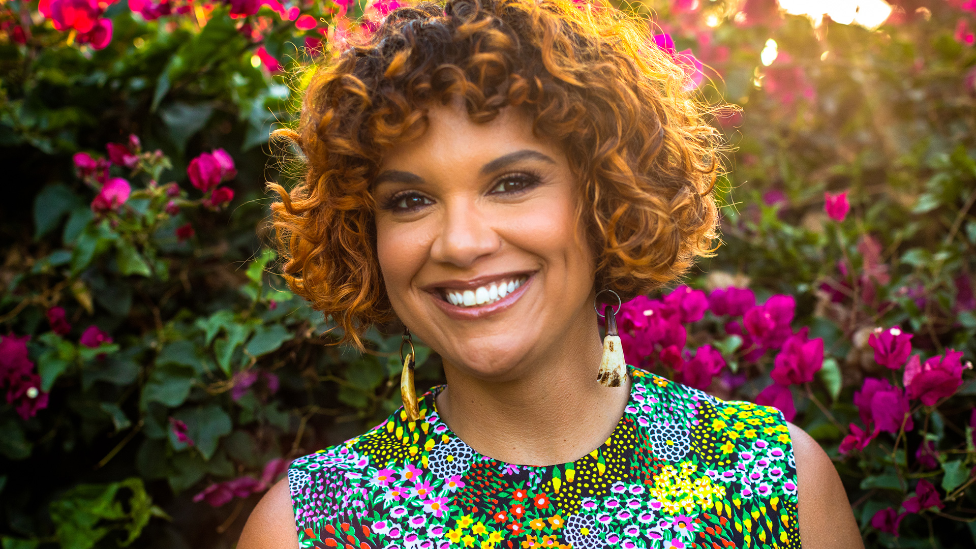 A vibrant portrait of Affrilachina poet and "Black by God" founder Crystal Good against pink bougainvillea flowers. Her curly hair is honey-colored curly hair and she wears a colorful floral print sleeveless dress, radiating warmth with her bright smile and statement earrings.