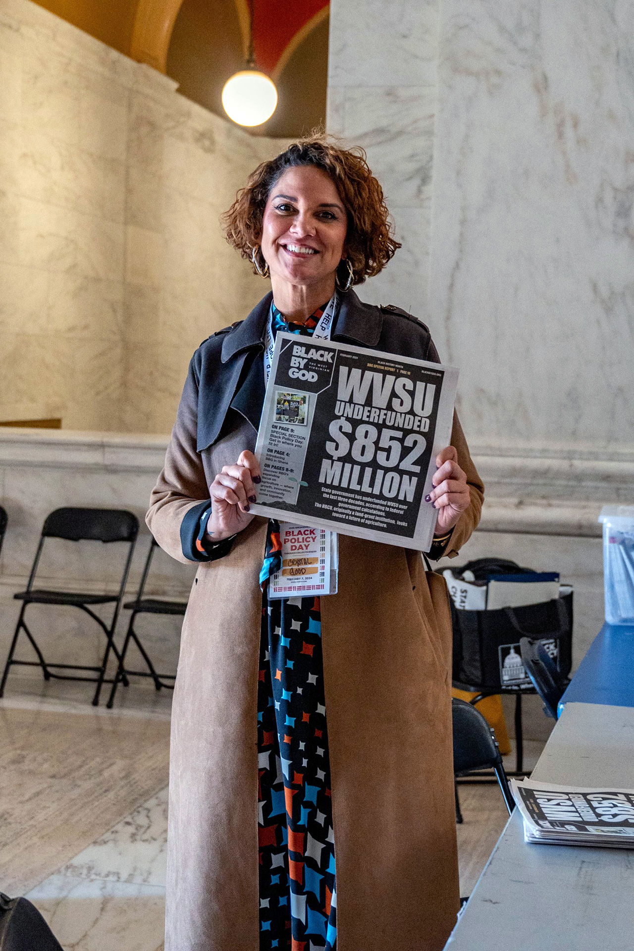 Crystal Good stands in a marble-walled hallway, proudly displaying the latest issue of the Black By God newspaper featuring a headline about West Virginia State University's $852 million underfunding.