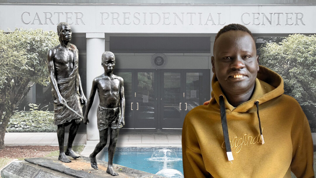 A South Sudanese refugee wearing a yellow hoodie smiles at the camera while standing in front of the Carter Presidential Center entrance. Behind him are two bronze statues depicting African figures, and a small fountain is visible in the background. The Center’s name is prominently displayed in large letters above the entrance.