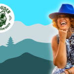 Affrilachian poet Crystal Good, founder of Black by God West Virginia, laughs joyfully wearing a bright blue hat and floral dress against an Appalachian mountain backdrop, embodying her mission to amplify Black journalism in West Virginia.