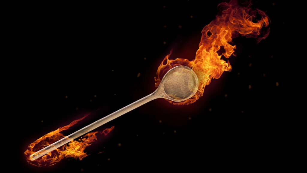 The image shows a dramatic artistic photograph of a wooden spoon engulfed in orange and red flames against a black background, with fire trailing from both the bowl and handle of the spoon. The composition symbolically represents the intersection of chronic illness spoon theory, trauma, and Appalachian wooden spoons through its powerful visualization of a kitchen implement transformed into something both destructive and beautiful.