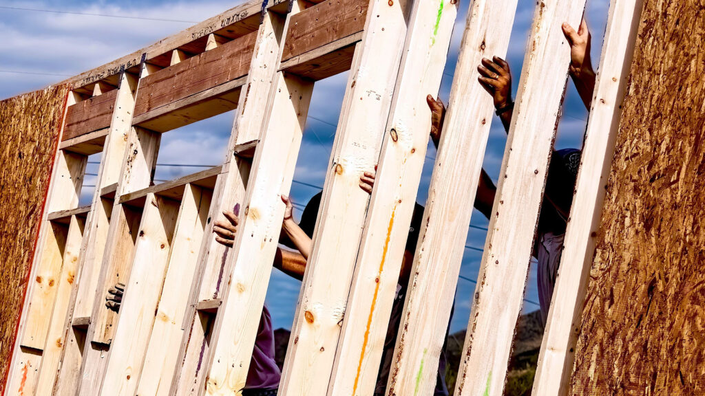 Hands holding vertical wooden studs as they raise the wall of a new house, against a blue sky background, with light-colored lumber arranged in a parallel pattern. Multiple pairs of hands are visible gripping the wooden beams.