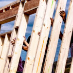 Hands holding vertical wooden studs as they raise the wall of a new house, against a blue sky background, with light-colored lumber arranged in a parallel pattern. Multiple pairs of hands are visible gripping the wooden beams.