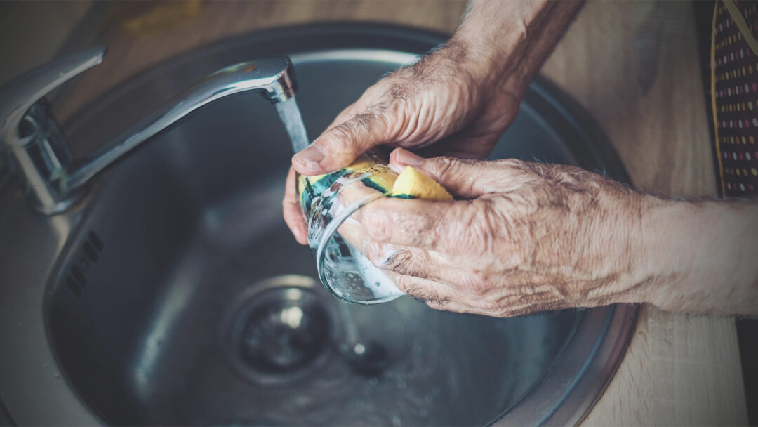 Weathered hands wash dishes at a sink, with strong fingers gripping a sponge and glass under running water. The intimate close-up captures the raw authenticity of Lucinda Zoe's Appalachian poetry, echoing mountain family stories through the daily rituals that define faith up in coal country.