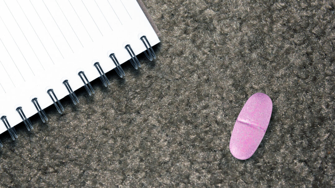 A pink pill on a carpet beside a spiral notebook, representing themes of the opioid crisis in rural Kentucky, Appalachian healthcare challenges, and the opioid epidemic in rural medicine.